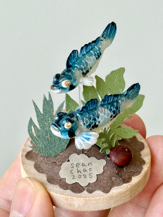 Small wooden ornament with two blue fish, leaves, and a small stone, held by a hand against a light gray background.