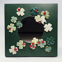 A square dark green panel with a circular mirror in the middle, surrounded by raised ceramic shamrock flowers in white, pale green, and teal, each accented with a small red ladybugs.