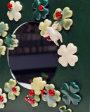 A square dark green panel with a circular mirror in the middle, surrounded by raised ceramic shamrock flowers in white, pale green, and teal, each accented with a small red ladybugs.