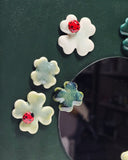 A square dark green panel with a circular mirror in the middle, surrounded by raised ceramic shamrock flowers in white, pale green, and teal, each accented with a small red ladybugs.