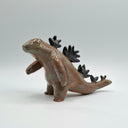 A small sculptural dinosaur figurine with a brown and green glazed body and black spikes along its back, photographed on a clean light gray studio background.