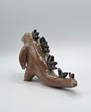 A glazed ceramic dinosaur figurine in a side profile, featuring a brown and green earthy glaze with black spikes along its back, set against a plain light gray background.