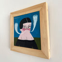 A framed painting featuring a stylized illustration of a woman with black hair, a pink face, and two blue ghostly figures coming out of her mouth, shaped like snakes on a blue background, housed in a light wood frame.