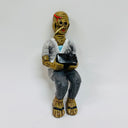 Clay sculpture of a very old man, depicted in an over the top way with brown, wrinkly skin and recessed eyes and nose - like a mummy. He sits and holds a black box.