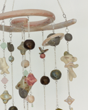 A hanging decorative mobile made from ceramic mini sculpts, small natural stones and bead charms on silver chains, photographed against a plain gray background.