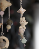 A hanging decorative mobile made from ceramic mini sculpts, small natural stones and bead charms on silver chains, photographed against a plain gray background.