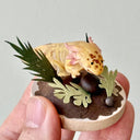 Sculpture of a clay axolotl, yellow in color, atop a wooden base with paper plants and small rocks.