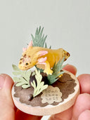 Sculpture of a clay axolotl, yellow in color, atop a wooden base with paper plants and small rocks.