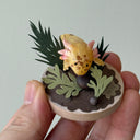 Sculpture of a clay axolotl, yellow in color, atop a wooden base with paper plants and small rocks.