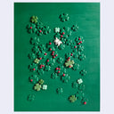 A green rectangular panel covered with scattered shamrock and four-leaf clover cutouts, plus a few small red ladybug accents and one white clover piece, photographed against a light gray background.