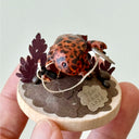 Sculpture of a orange and brown striped salamander standing on a wooden base and holding a kusarigama.