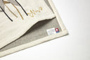A close-up of a beige linen textile showing embroidered details of a signature and gold script, with a gray inner lining and a small sewn-in care label.