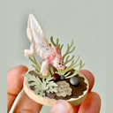 Sculpture of a clay axolotl, pink in color, atop a wooden base with paper plants and small rocks.
