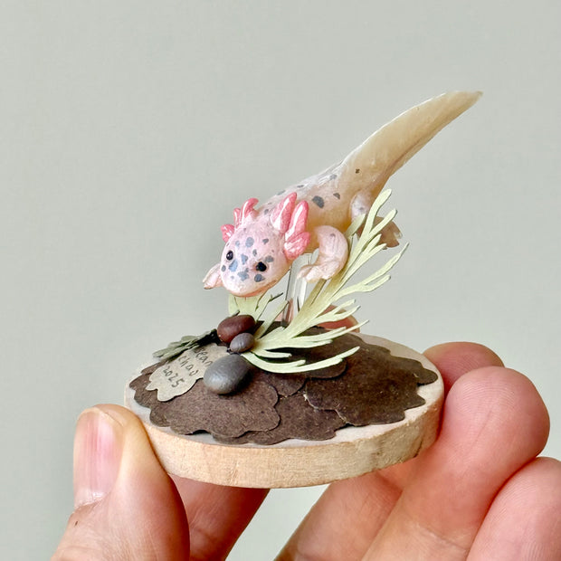 Sculpture of a clay axolotl, pink in color, atop a wooden base with paper plants and small rocks.