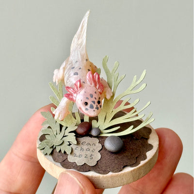 Sculpture of a clay axolotl, pink in color, atop a wooden base with paper plants and small rocks.