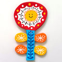Flat, colorful clay sculpture of a red and white flower with a blue patterned stem and orange and yellow leaves. It has a smiley face as the yellow flower center.