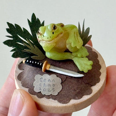 Small sculpture of a frog on a wooden base with leaves and a small sword, held by a hand.