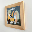 A framed whimsical illustration of two stylized figures with large beige hairdos, one seated in a black outfit and one standing in a white dress, set against a dark blue-gray background. The artwork is housed in a light natural wood frame and mounted on a white wall.