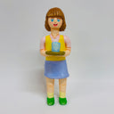 Clay sculpture of a woman with short brown hair with bangs, smiling and holding a tray with a cup of tea on it. She wears a pink shirt, yellow vest and blue skirt.