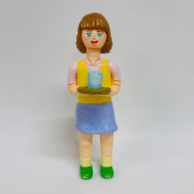 Clay sculpture of a woman with short brown hair with bangs, smiling and holding a tray with a cup of tea on it. She wears a pink shirt, yellow vest and blue skirt.
