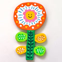 Flat clay sculpture of a flower, orange and white with blue and yellow color accents. It has a smiley face in its center and is attached to a green stem.