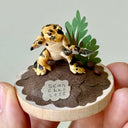 Small figurine of a leopard colored newt holding a shurikan on a wooden base with plants and rocks, held by a hand.