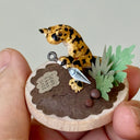 Small figurine of a leopard colored newt holding a shurikan on a wooden base with plants and rocks, held by a hand.