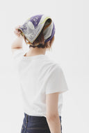 Person wearing a white t-shirt and blue pants with a purple and patterned headscarf on a plain background