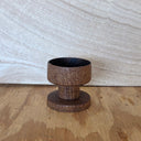 A small dark brown wooden pedestal cup with a circular base, shown on a wooden tabletop against a light textured wall.