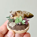 Sculpture of a clay axolotl, brown in color, atop a wooden base with paper plants and small rocks.