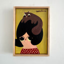A framed hand-painted folk artwork featuring a stylized woman with black hair, a brown squirrel on her head, and a red polka-dot top against a mustard yellow background, mounted on a white wall. Both the girl and the squirrel smoke a cigarette.