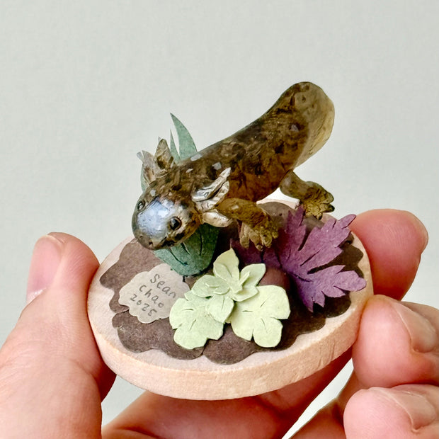Sculpture of a clay axolotl, brown in color, atop a wooden base with paper plants and small rocks.
