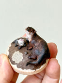 Small sculpture of 2 otters - a larger one and a baby - on a wooden base covered in paper with small rocks.