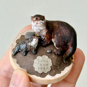 Small sculpture of 2 otters - a larger one and a baby - on a wooden base covered in paper with small rocks.
