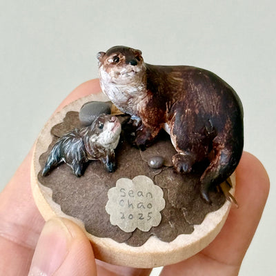 Small sculpture of 2 otters - a larger one and a baby - on a wooden base covered in paper with small rocks.