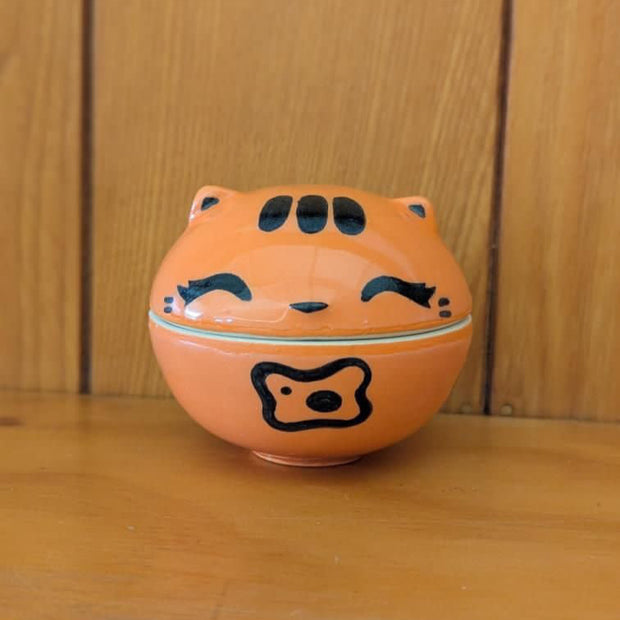 Ceramic sculpture of an orange cat jar, with a shallow bowl for the bottom and a cat head for the lid. The cat has simply drawn patterning, ears and closed eyes with thick eyeliner.