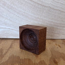 A small square dark wood block with a carved spiral standing on a wooden surface against a light wood background.