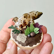 Sculpture of a clay axolotl, brown in color, atop a wooden base with paper plants and small rocks.