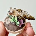 Sculpture of a clay axolotl, brown in color, atop a wooden base with paper plants and small rocks.
