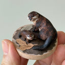 Small sculpture of 2 otters - a larger one and a baby - on a wooden base covered in paper with small leaves.