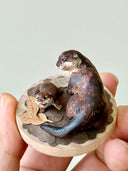 Small sculpture of 2 otters - a larger one and a baby - on a wooden base covered in paper with small leaves.