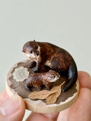 Small sculpture of 2 otters - a larger one and a baby - on a wooden base covered in paper with small leaves.