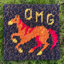 Cross-stitched orange horse with a red mane and 'OMG' text on a dark background, placed on grass.