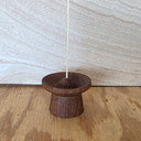 A small dark brown wooden incense holder with a conical top and a single incense stick inserted, set on a wooden surface against a light wood-paneled background.
