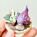 Sculpture of a clay axolotl, purple in color, atop a wooden base with paper plants and small rocks.
