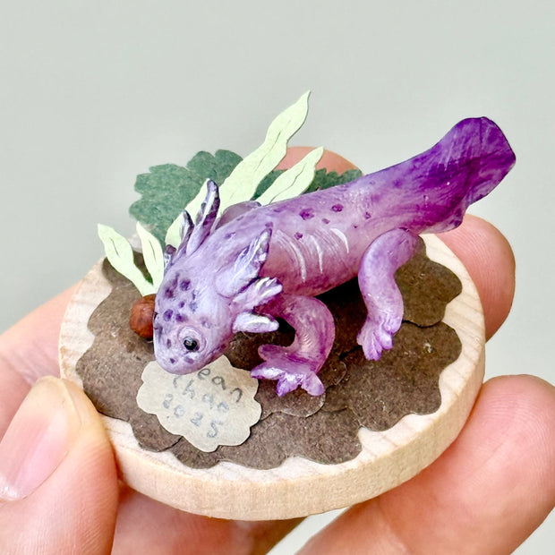 Sculpture of a clay axolotl, purple in color, atop a wooden base with paper plants and small rocks.