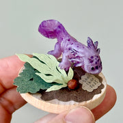 Sculpture of a clay axolotl, purple in color, atop a wooden base with paper plants and small rocks.