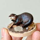 Small sculpture of 2 otters - a larger one and a baby - on a wooden base covered in paper with small leaves.