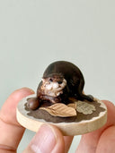 Small sculpture of 2 otters - a larger one and a baby - on a wooden base covered in paper with small leaves.