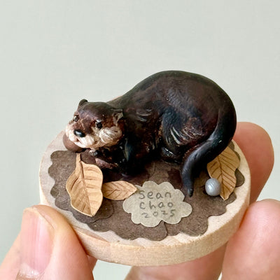Small figurine of an otter on a round wooden base with leaves and a pearl, held by a hand against a light background.